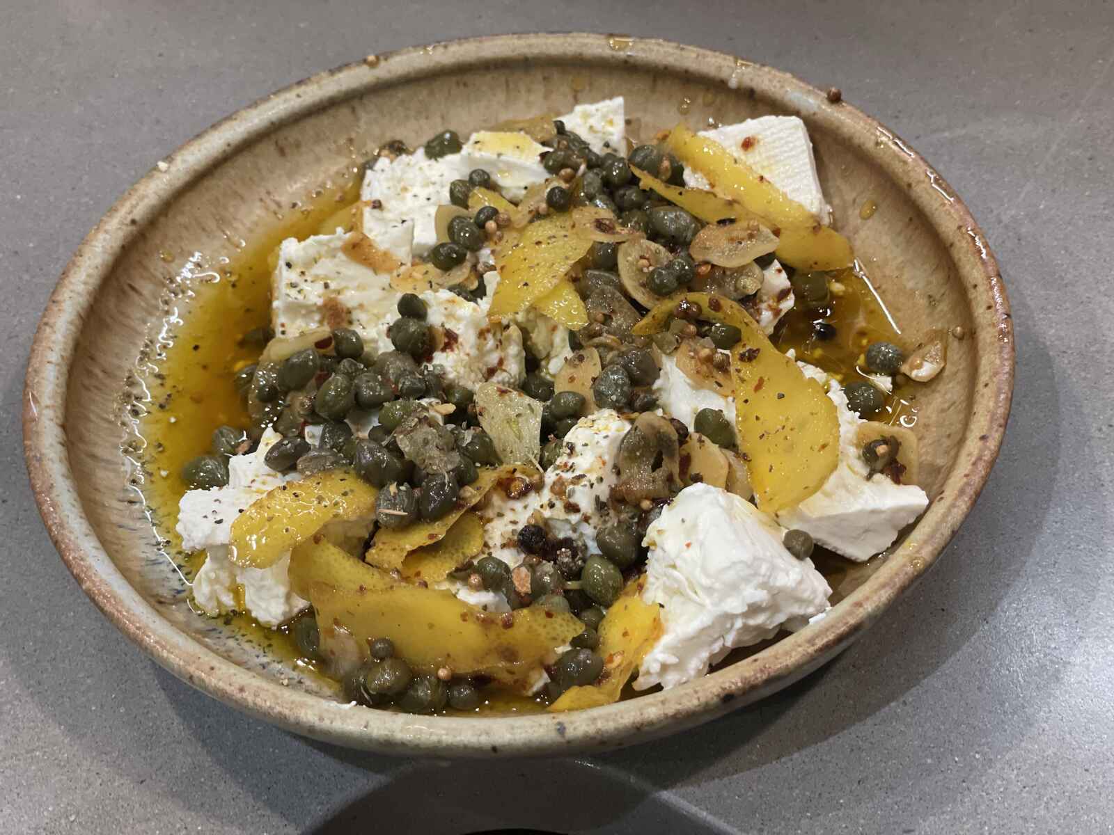 A shallow bowl containing feta cheese with lemon peel, capers, garlic, cumin, coriander, red chili flake and olive oil on top