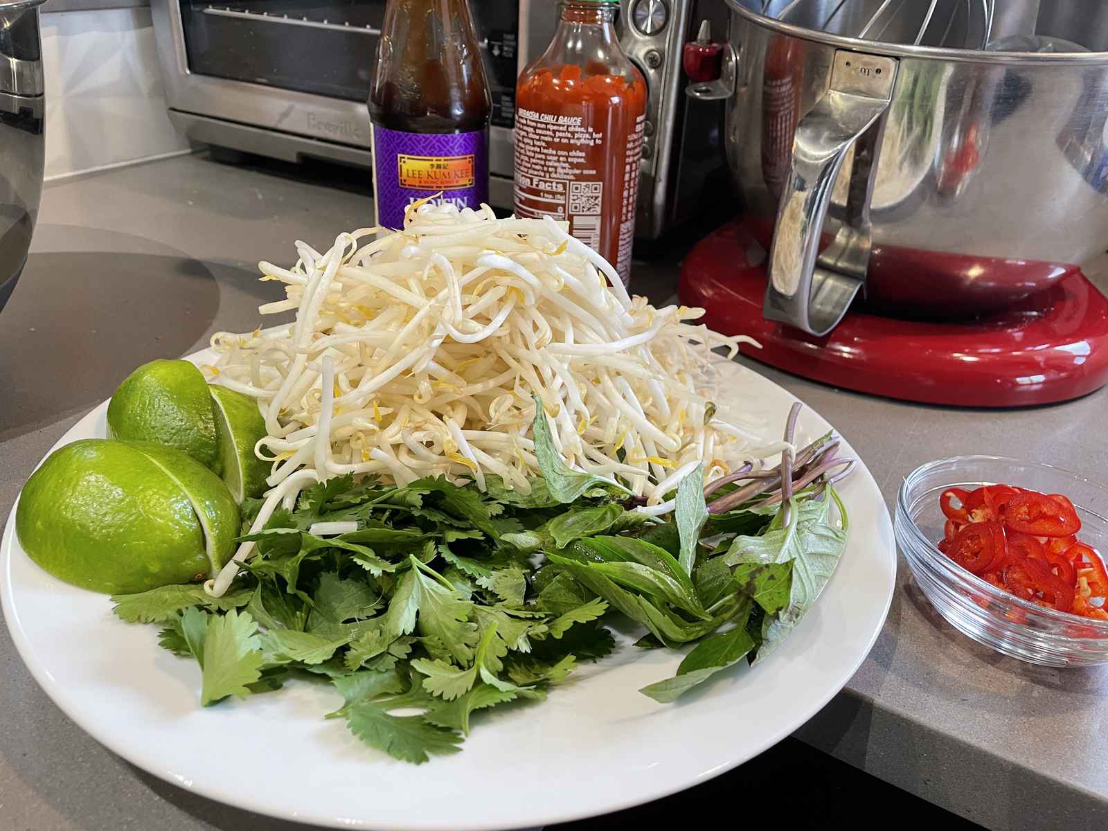 A pile of bean sprouts, basil, cilantro and limes along with hosin and sriracha